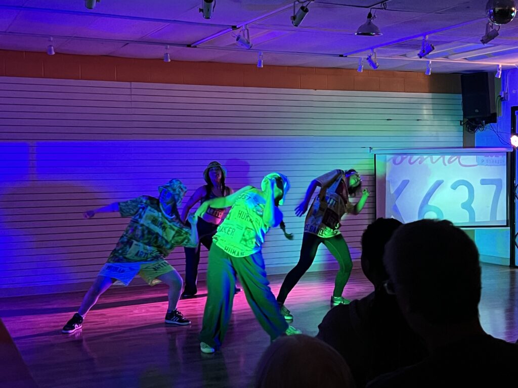 Four dancers lit in purple and green, leanin to the right. The dancers wear clothing with license plate-like images reading "HUMAN ARTIST." In the background, a projector shows an image of a license plate.